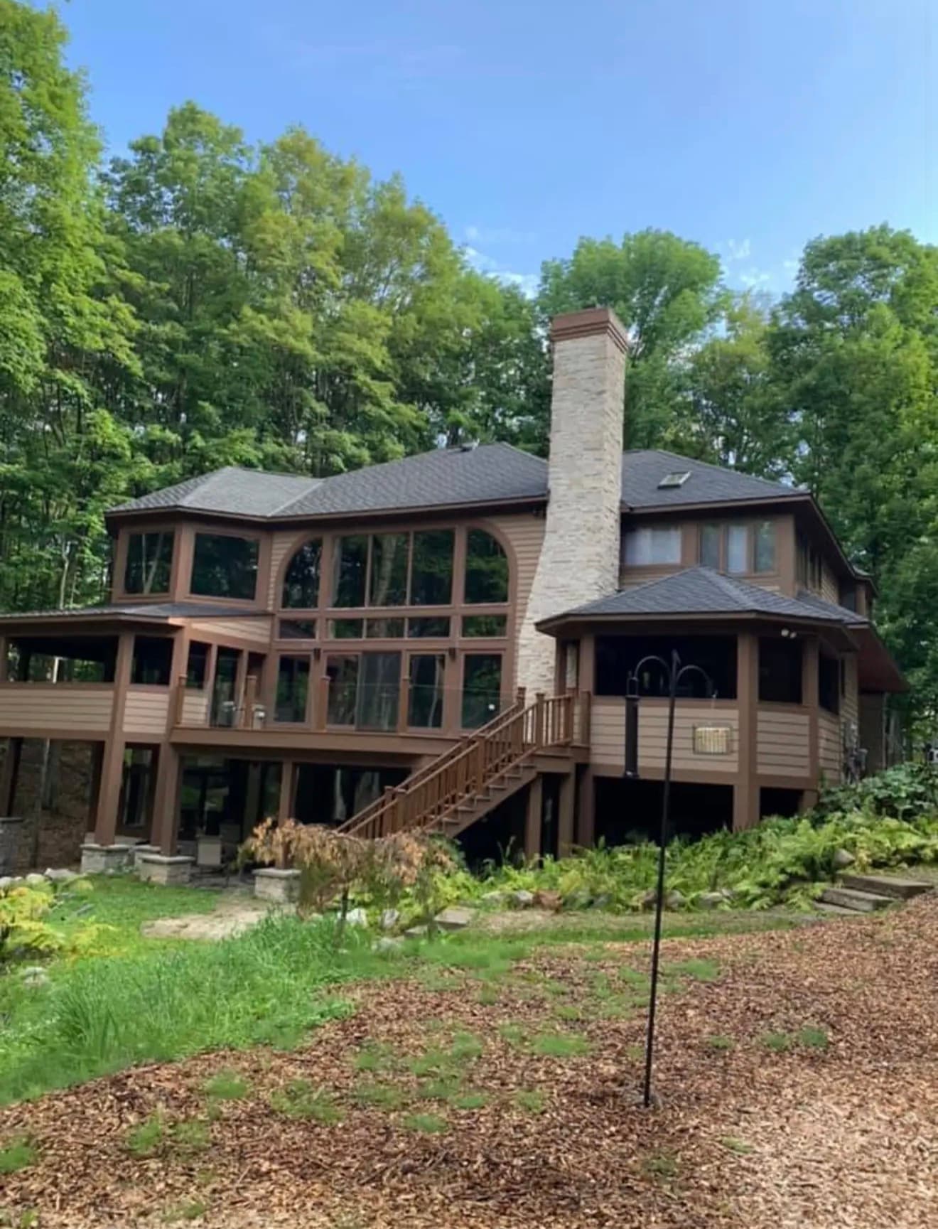 Completed lakeside craftsman home in a Northern Michigan forest, multi-level wraparound deck and a stone chimney rising through the architectural shingle roof